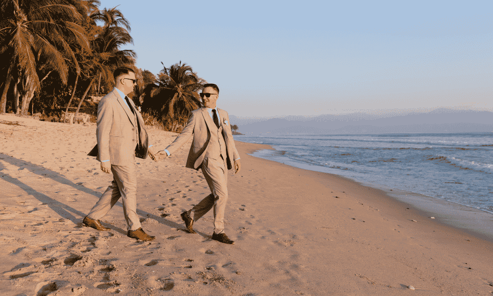 A newly married couple in their wedding outfits walking hand in hand on the beach.